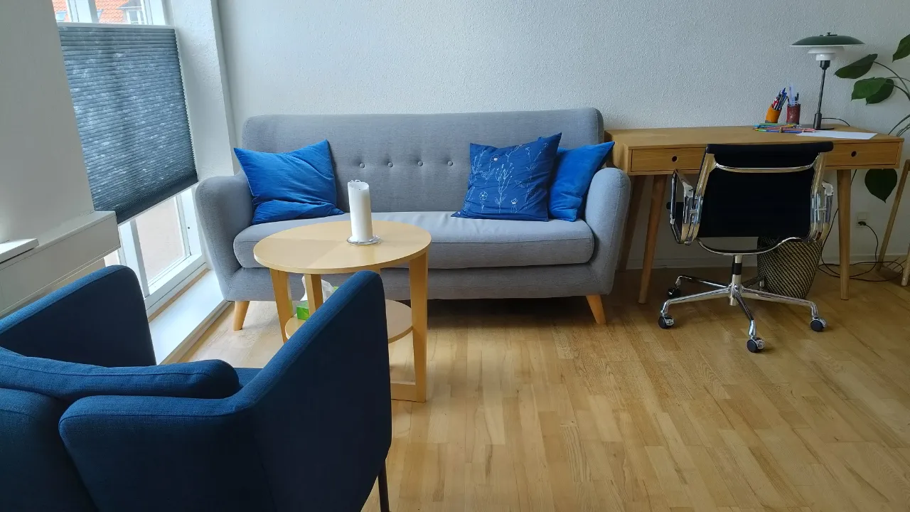 Moderne stue med grå sofa, blå puder, træbord, skrivebord og kontorstol ved stort vindue.