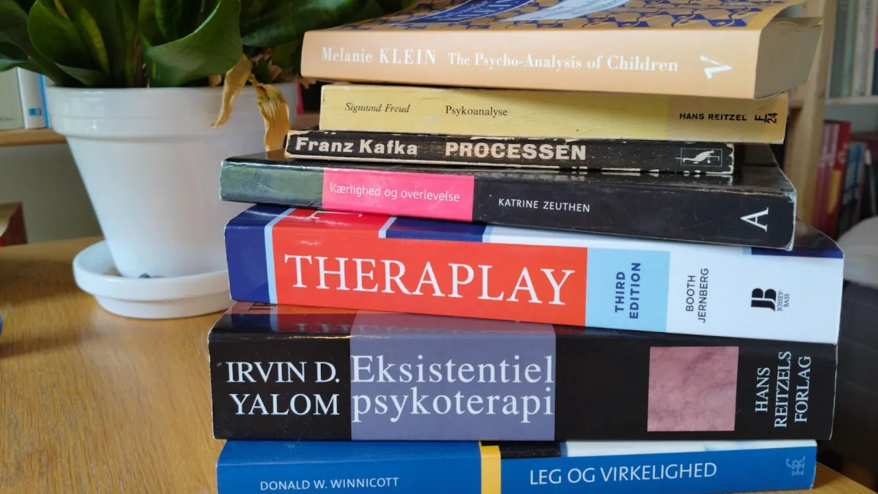 Stak bøger om psykoterapi, psykoanalyse og filosofi på træbord ved siden af en hvid plantekrukke.