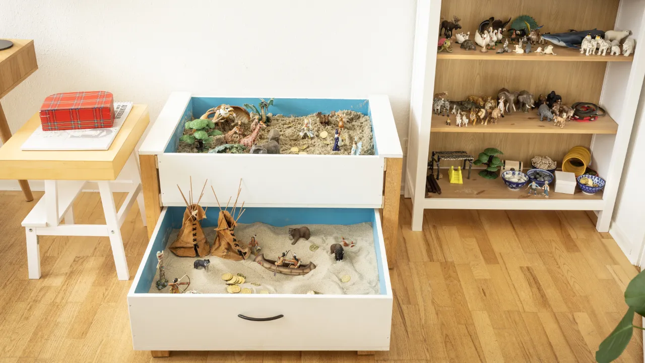 Legetøjslandskaber med sand og miniaturefigurer i to hvide skuffer, ved siden af reol med figurer og rekvisitter.